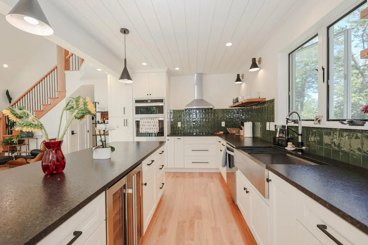 Professionally staged white kitchen with a long island, black counters, green tile, and forest-facing window at Grateful Ledge